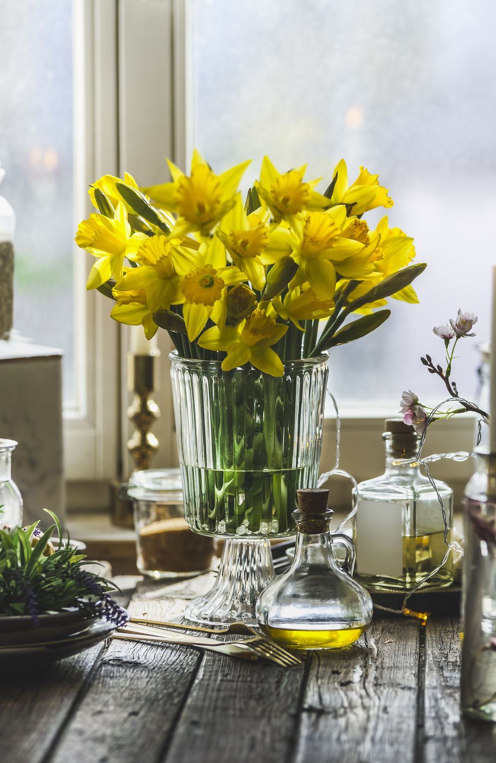 yellow daffodils flowers bunch in glass vase on kitchen table with candles , plates , jars and bottles at window background cozy springtime at home still life
