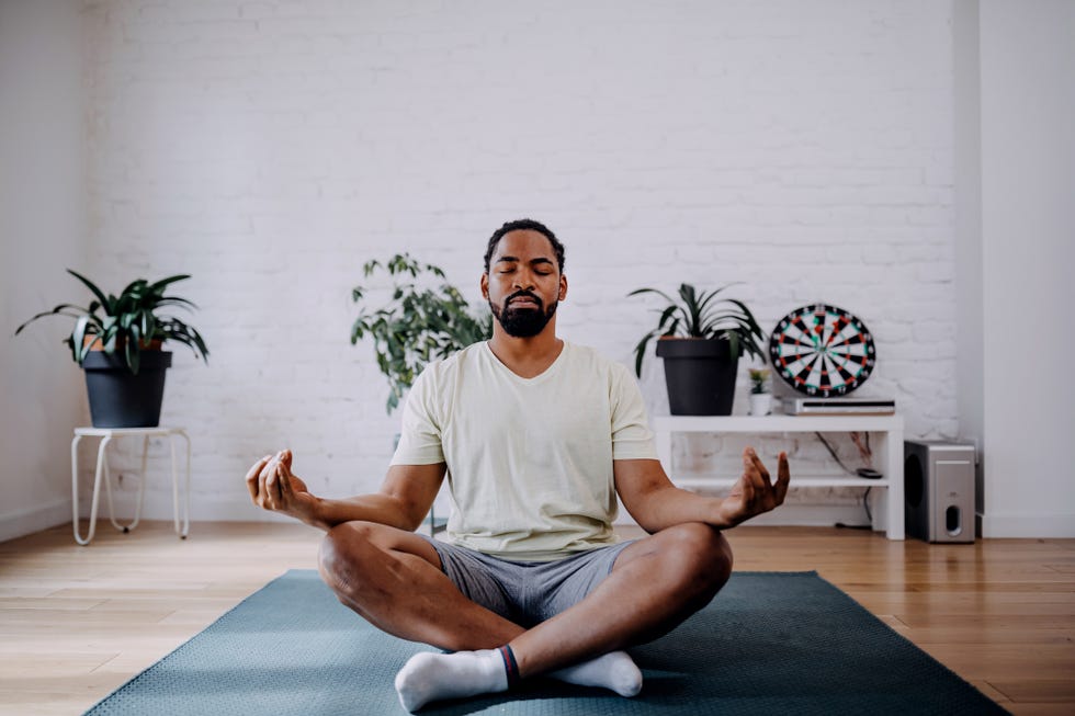 sporty mid adult black man meditating alone at home, peaceful calm hipster fit guy practicing yoga in lotus pose indoors holding hands in mudra, freedom and calmness concept