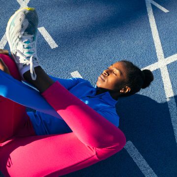 a black afro american girl is on the floor performing stretches on the blue running track she is wearing pink leggings and a blue sweater