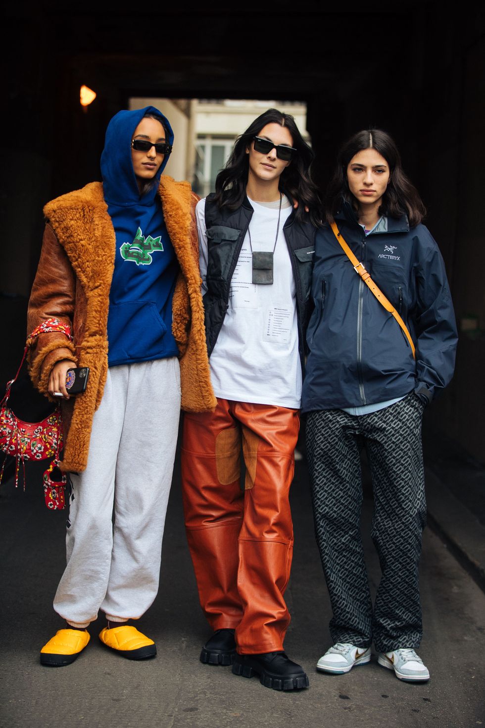 paris, france march 02 models mona tougaard, vittoria ceretti, and loli bahia after the courrèges show at garage amelot during paris fashion week fallwinter 2022 on march 02, 2022 in paris, france mona wears an oversized brown shaggy shearling jacket, blue hoodie, white trousers, red floral balenciaga print purse, yellow north face down slippers vittoria wears a black vest, small black neck pouch, white mm6 maison margiela shirt, brown leather pants, and black chunky boots loli wears a blue arcteryx jacket, black chain print pants, and gray and white nike dunk sneakers photo by melodie jenggetty images