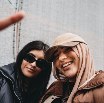 two young adult girls are making funny gestures to the camera one of them is wearing a fashionable hijab with a hat