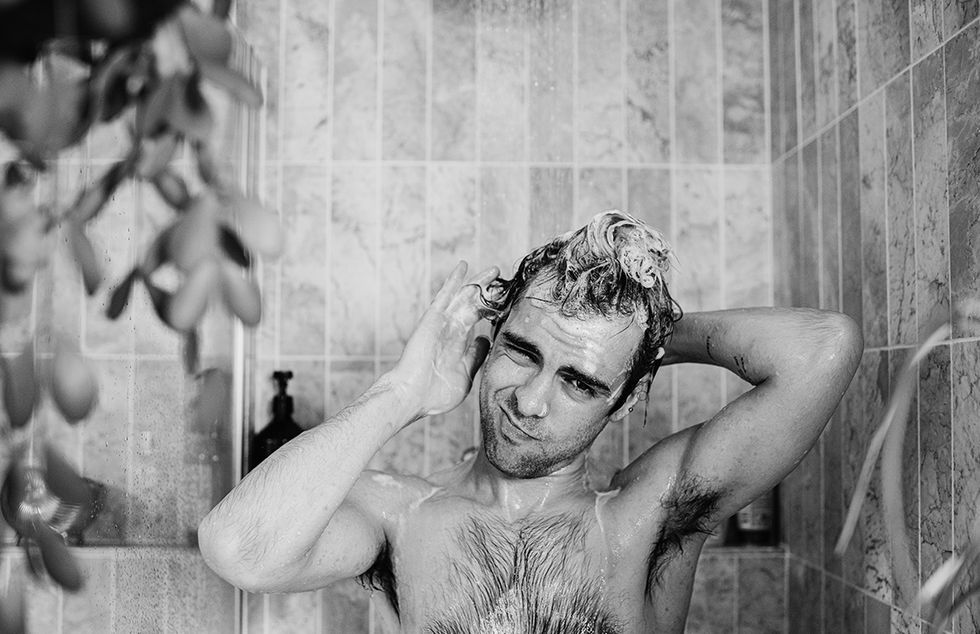 a man stands in the shower, scrubbing his head and creating a lather with the shampoo he looks at the camera as he washes behind his ear