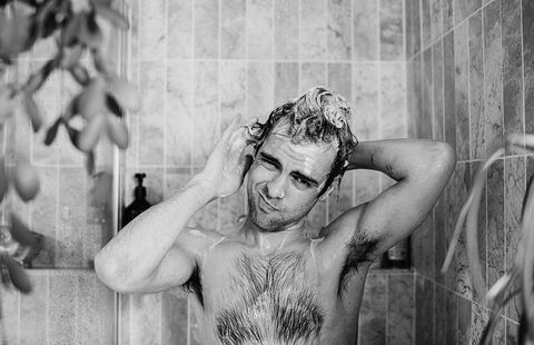 a man stands in the shower, scrubbing his head and creating a lather with the shampoo he looks at the camera as he washes behind his ear