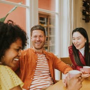 Three young people sit around a table and giggle as they have a lighthearted discussion and drink coffee / tea three young people socialise at a table while enjoying hot drinks they are animated as they giggle together one girl is in the foreground whilst focus remains on a smiling male and female