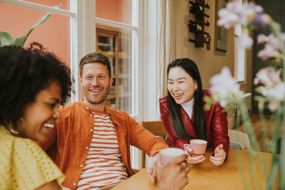 three beautiful young people socialise at a table while enjoying hot drinks they are animated as they giggle together one girl is in the foreground whilst focus remains on a smiling male and female