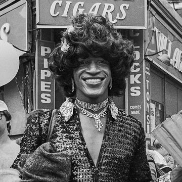 marsha p johnson in dark outfit and black hair smiles as she stands on the corner of christopher street and 7th avenue during the pride march