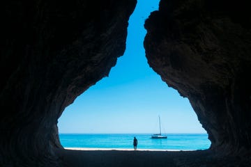 a sailboat by the coastline in a turquoise sea, framed by the silhouetted cliffs of a cave, with a silhouetted person on the beach, in landscape format