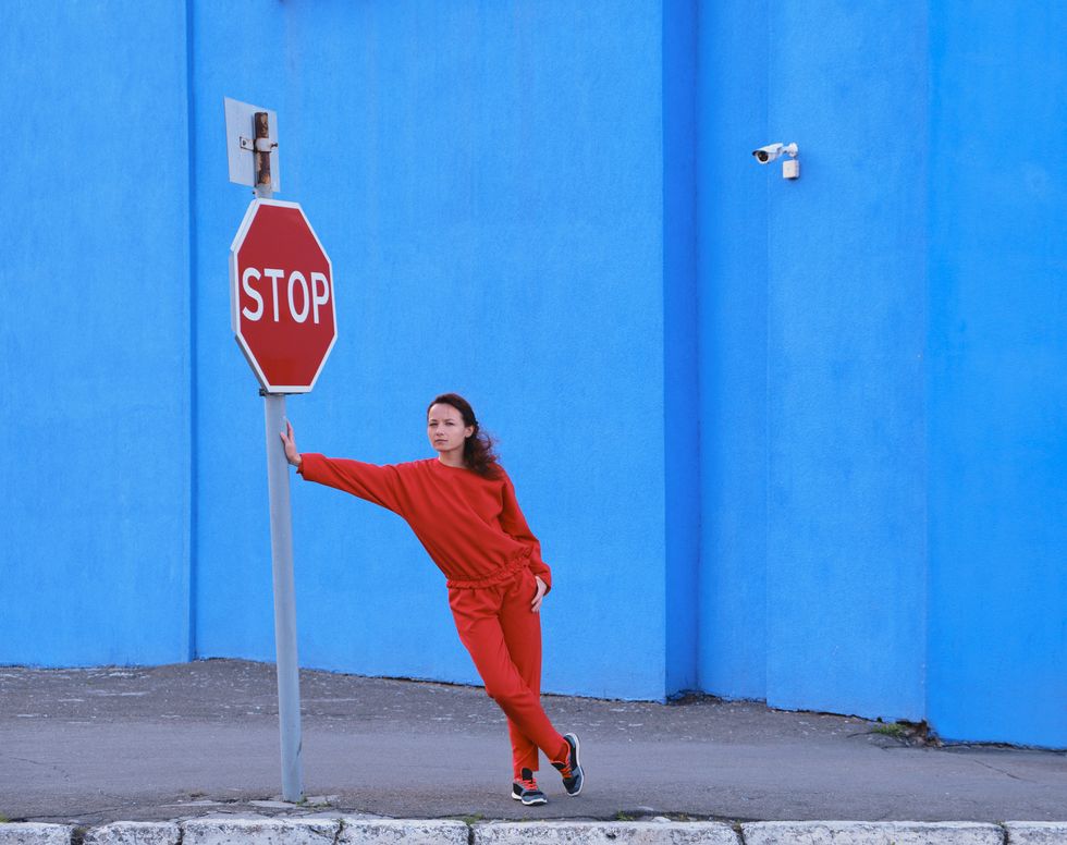 female, dressed in red casual costume, leans her hand on the road sign stop looking in left side of frame vibrant blue wall background color contrast of red blue colors concept idea for waiting , patience, expectation