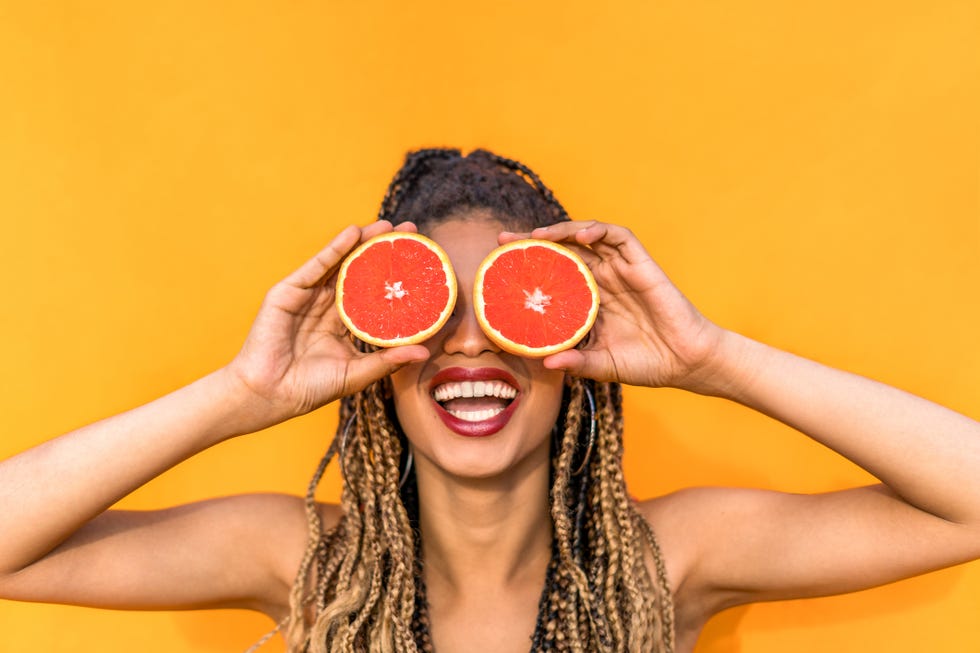 smiling woman holding blood orange sclices ,fruits covering her eyes on orange background