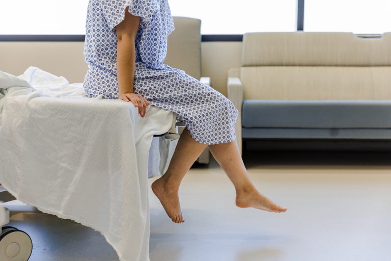 an unrecognizable female patient wears a hospital gown and sits on the edge of the hospital bed. she dangles her feet while waiting for her test results.