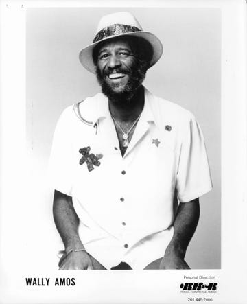 wally amos also known as famous amos smiles at the camera, he wears a collared shirt and hat