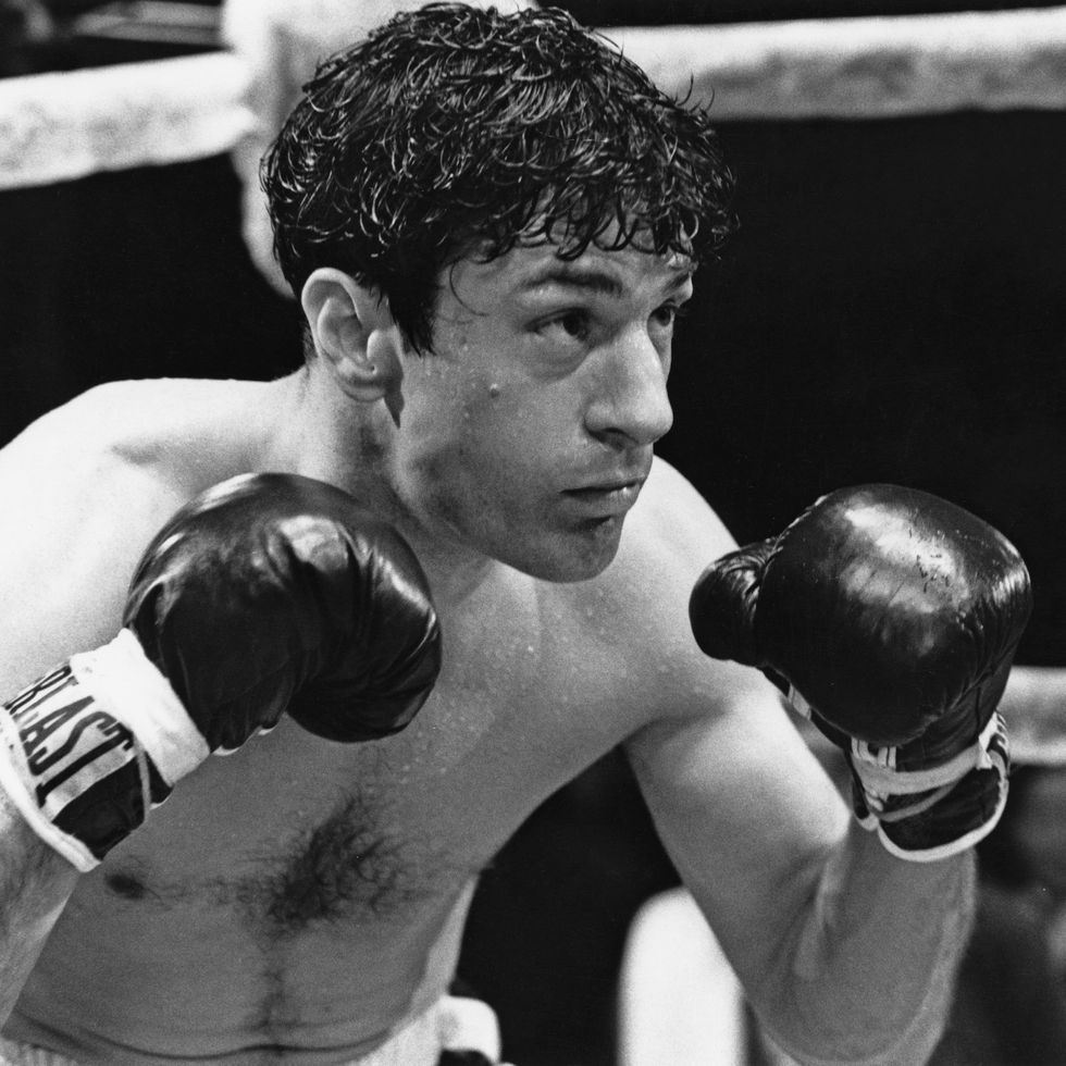 robert de niro in character as jake lamotta in raging bull, he holds both of his gloved fists up in a boxing ring and is shirtless