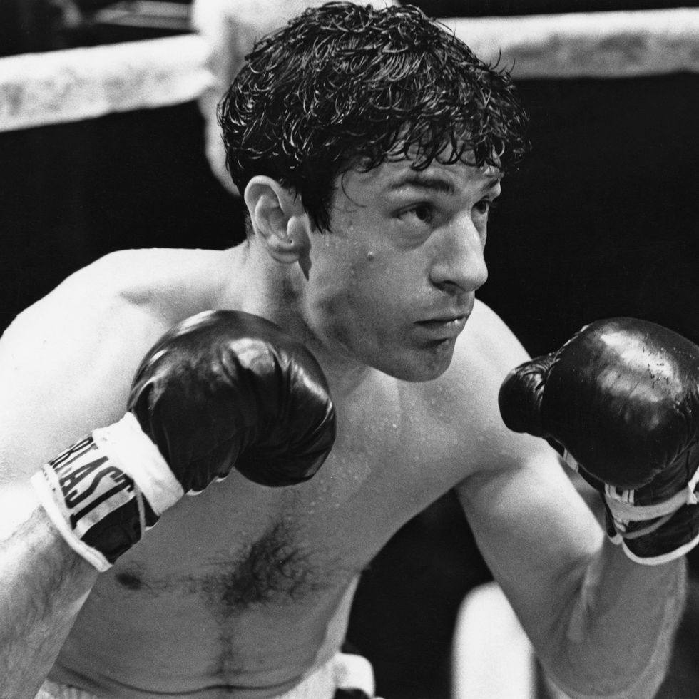 robert de niro in character as jake lamotta in raging bull, he holds both of his gloved fists up in a boxing ring and is shirtless