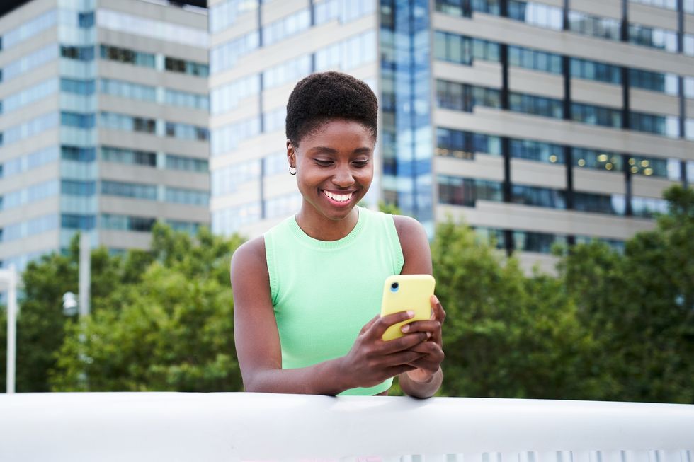 portrait of an african woman smiling while using phone outdoors to be connected, chatting with people at the city concept of mobile app user