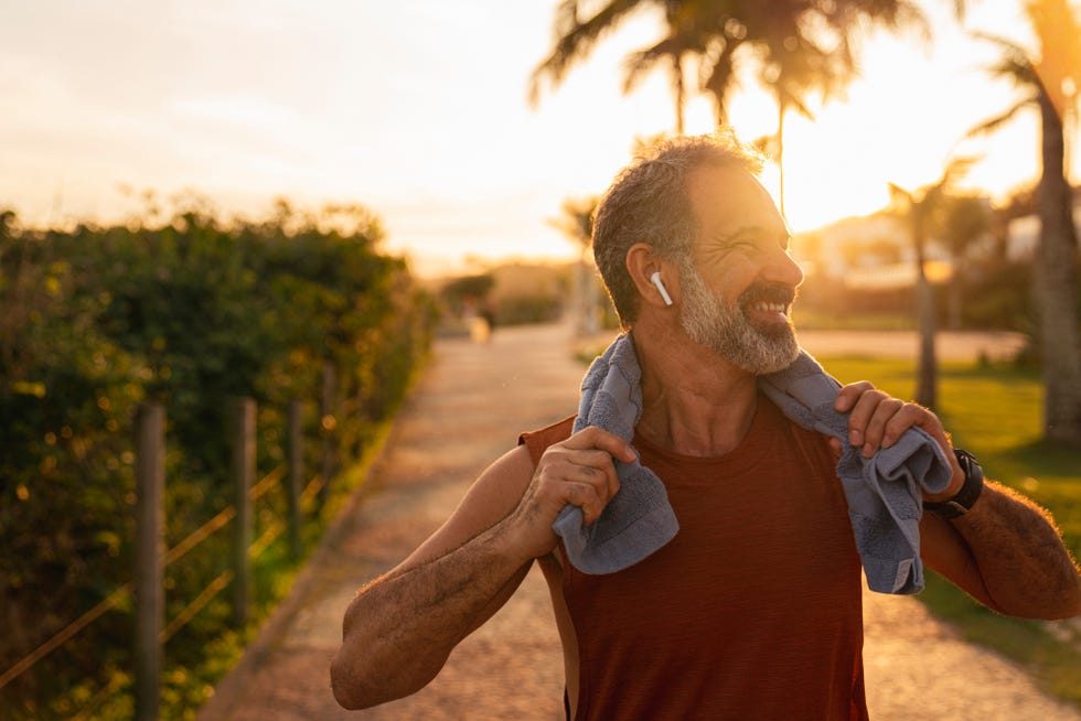 a motivated senior athlete, standing with a smartphone in his hand and a towel around his neck he catches his breath after running and rests
