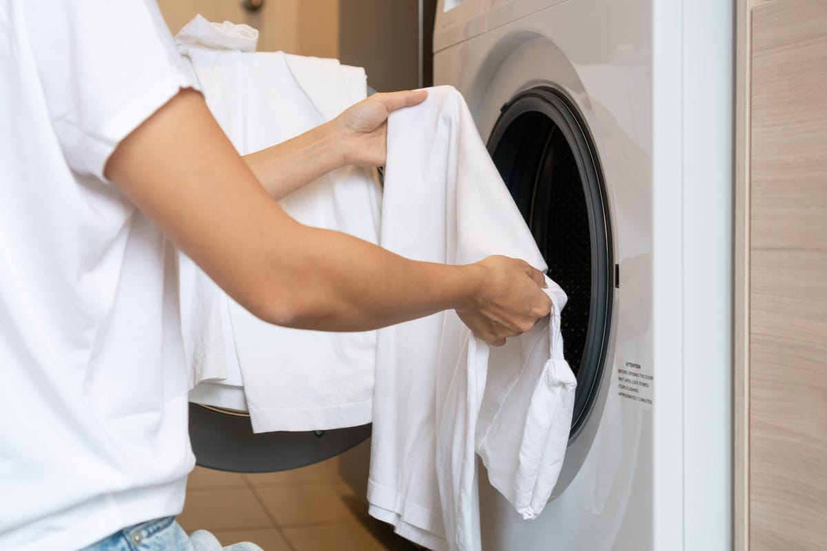 woman loading white color clothes into washing machine in kitchen at home. close up. laundry concept.