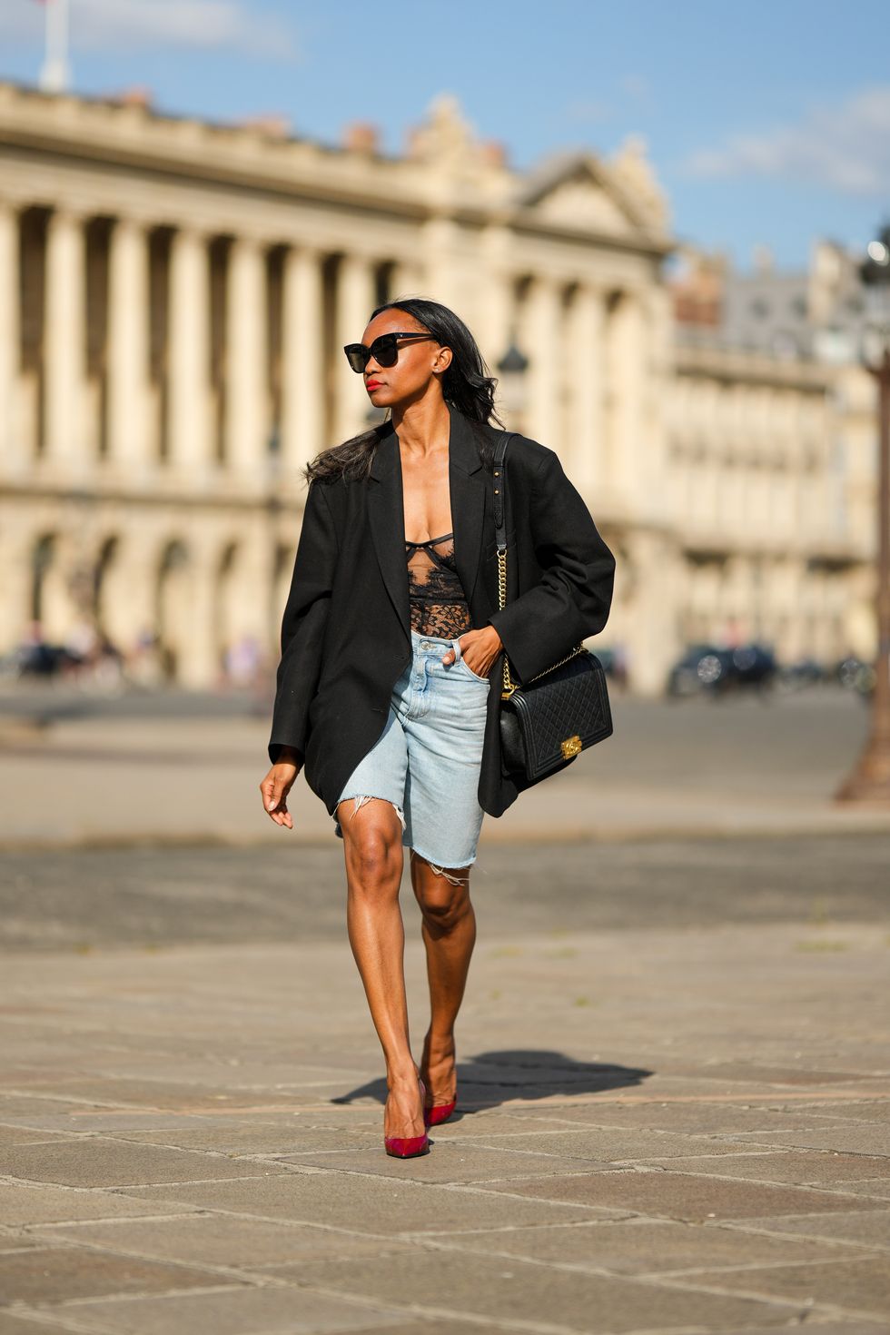 paris, france june 29 emilie joseph infashionwetrust wears black sunglasses, diamonds earrings, a black flower print pattern lace corset top, a black oversized blazer jacket from the frankieshop, blue denim ripped bermudas shorts, a black shiny leather large boy shoulder bag from chanel, gold bracelets, red shiny leather snake print pattern pointes pumps heels shoes from saint laurent paris, during a street style fashion photo session, on june 29, 2022 in paris, france photo by edward berthelotgetty images