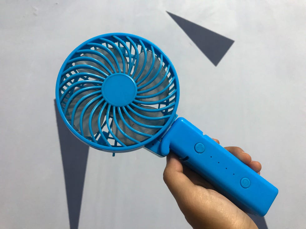 left palm of hand holding blue colored hand fan, mini fan with abstract background.