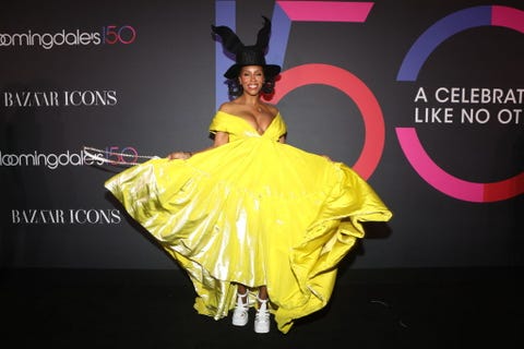 june ambrose on the red carpet in a yellow gown, white platform fisherman sandals, and a black hat with "bunny ears" shapes