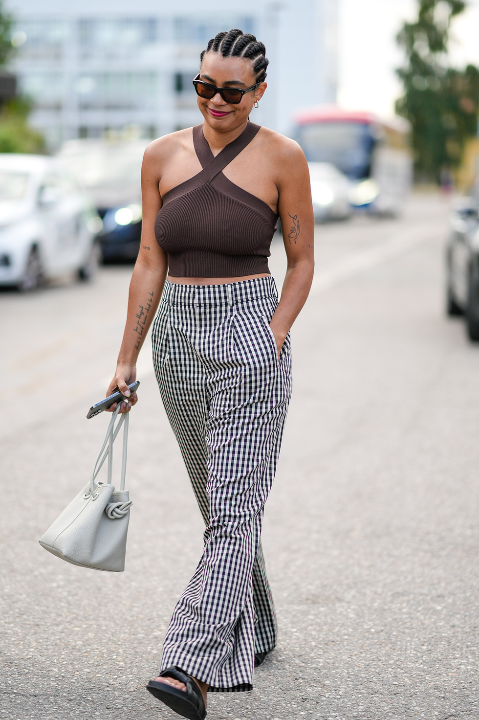 Kopenhagen, Denemarken, 9 augustus: een gast draagt een zwarte zonnebril, een donkerbruine haltertop/croptop, een zwart-witte wijde broek met klein ruitjesmotief, zwarte glanzende leren sandalen met klittenbandsluiting en bandjes, een witte handtas van mat latte leer, gouden ringen, gouden oorbellen, buiten Rabens Saloner, tijdens de Copenhagen Fashion Week lente/zomer 2023, op 9 augustus 2022 in Kopenhagen, Denemarken. (foto door Edward Berthelot/Getty Images)
