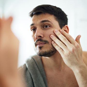 close up shot of handsome man with beard using anti aging moisturizer in the morning he is applying the cream under his eyes while looking at himself in the mirror