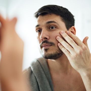 close up shot of handsome man with beard using anti aging moisturizer in the morning he is applying the cream under his eyes while looking at himself in the mirror