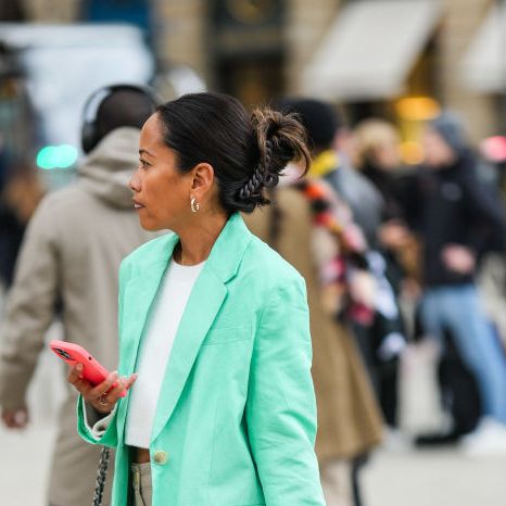 a guest wears a white cropped top, a mint green long blazer jacket, a black shiny leather handbag from chanel, beige large pants, a white latte and mint green suede sneakers, black sunglasses, a black hair clip , outside schiaparelli, during paris fashion week