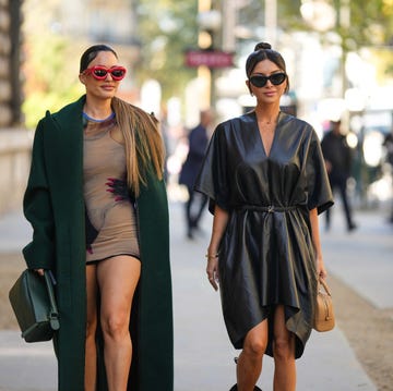 paris, france september 30 karen wazen wears red puffy loewe sunglasses, a beige and black body print pattern tube short skirt from loewe, a dark green oversized long coat, a dark green shiny leather puzzle handbag from loewe, , nathalie fanj wears black sunglasses, gold earrings, a gold chain necklace, a black shiny leather bat sleeves short dress from loewe, a black shiny leather with silver logo belt from loewe, a gold juste un clou bracelet from cartier, gold rings, a brown leather handbag from loewe, outside loewe, during paris fashion week womenswear springsummer 2023, on september 30, 2022 in paris, france photo by edward berthelotgetty images