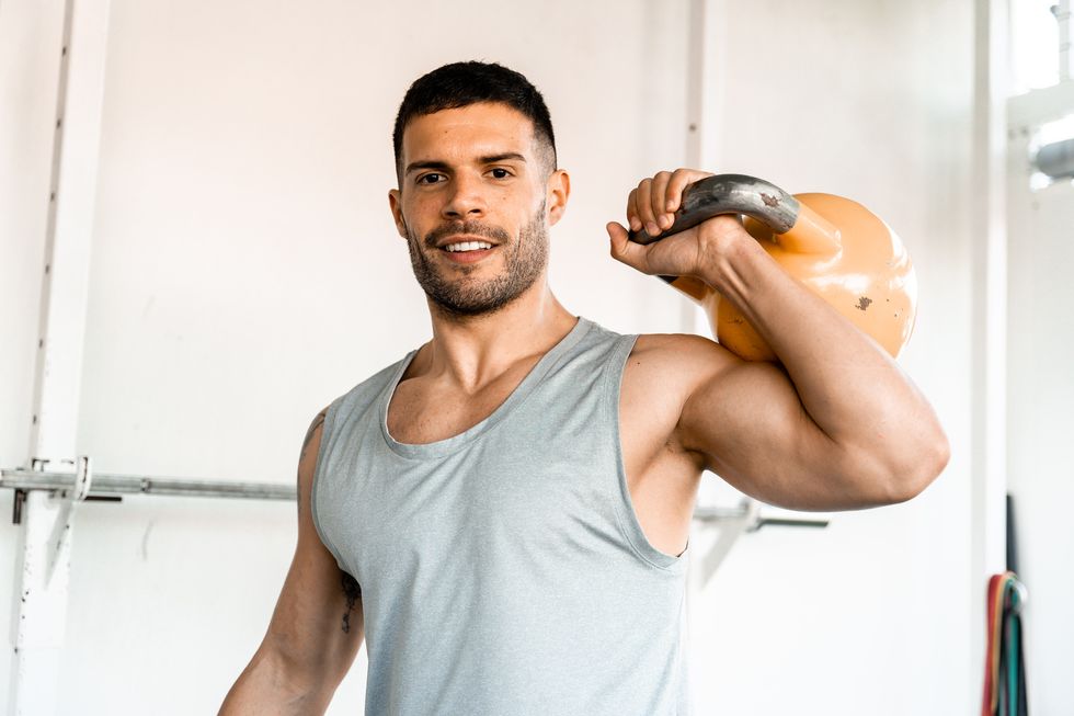 man smiling at the camera while holding kettlebell over his shoulder male athlete holding kettlebell doing gym workout in gym