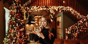 a smiling happy young woman looks at her phone communicates online using wireless technologies and talks on her smartphone celebrating the christmas holiday in a decorated interior with a christmas tree on a new year's holiday in winter