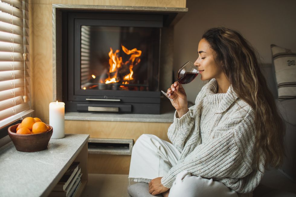 smiling woman enjoying alcohol drink at fireside wearing in warm woolen sweater romantic christmas evening, time to self beautiful light from burning fire and candle