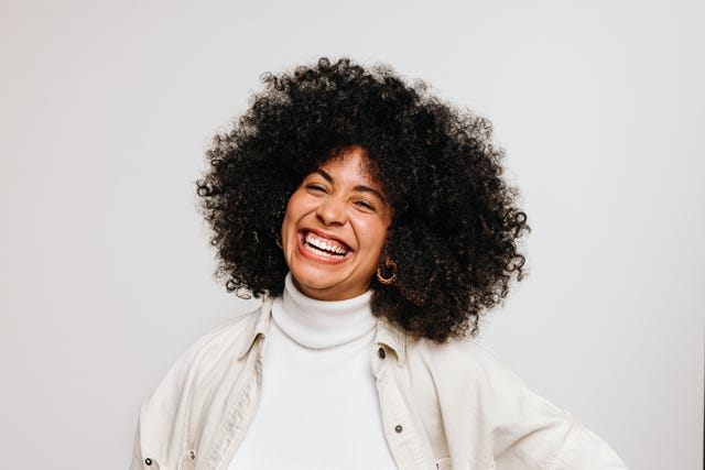 beautiful young woman with an afro hairstyle smiling at the camera while standing against a white background happy woman of color wearing her natural curly hair with pride