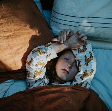 a cute young girl lies in a comfy double bed, wrapped in a thick duvet with a terracotta coloured linen duvet cover and white bedding her arms are raised above her head and over her forehead in a cute position
