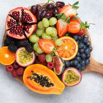 delicious fruit on round wood chopping board, mango pomegranate raspberries papaya oranges passion fruits berries on off white concrete background, selective focus