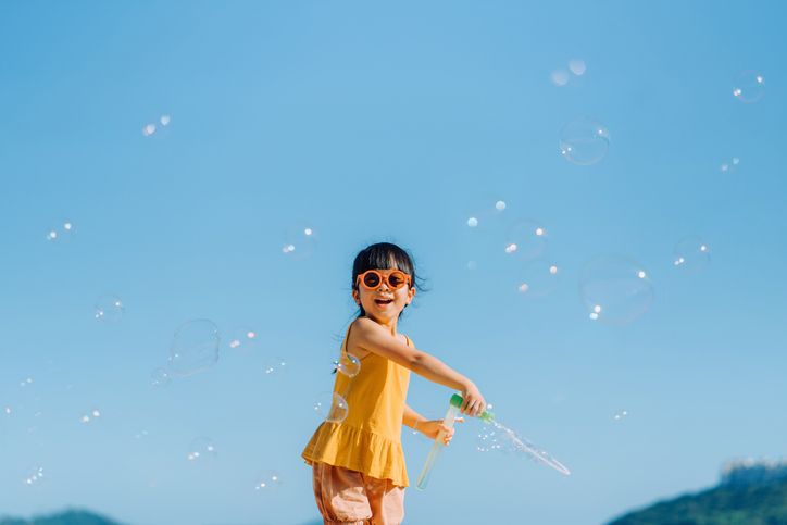 happy little asian girl with sunglasses having fun playing and blowing soap bubbles outdoors on a beautiful sunny day against blue sky