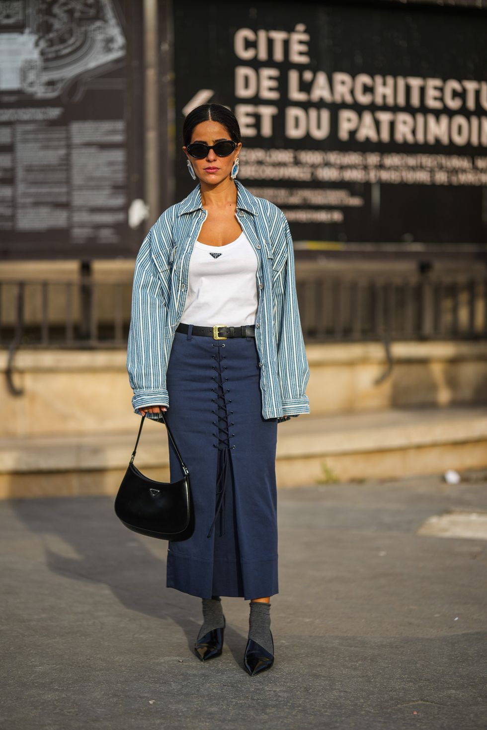 paris, france october 03 a guest wears black sunglasses, blue earrings, a white tank top from prada, a blue and white striped print pattern shirt, a black shiny leather belt, a navy blue laces front long skirt, a black shiny leather cleo handbag from prada, gray wool socks, black shiny varnished leather asymmetric heels mules from prada, outside rokh, paris fashion week womenswear springsummer 2023, on october 03, 2022 in paris, france photo by edward berthelotgetty images