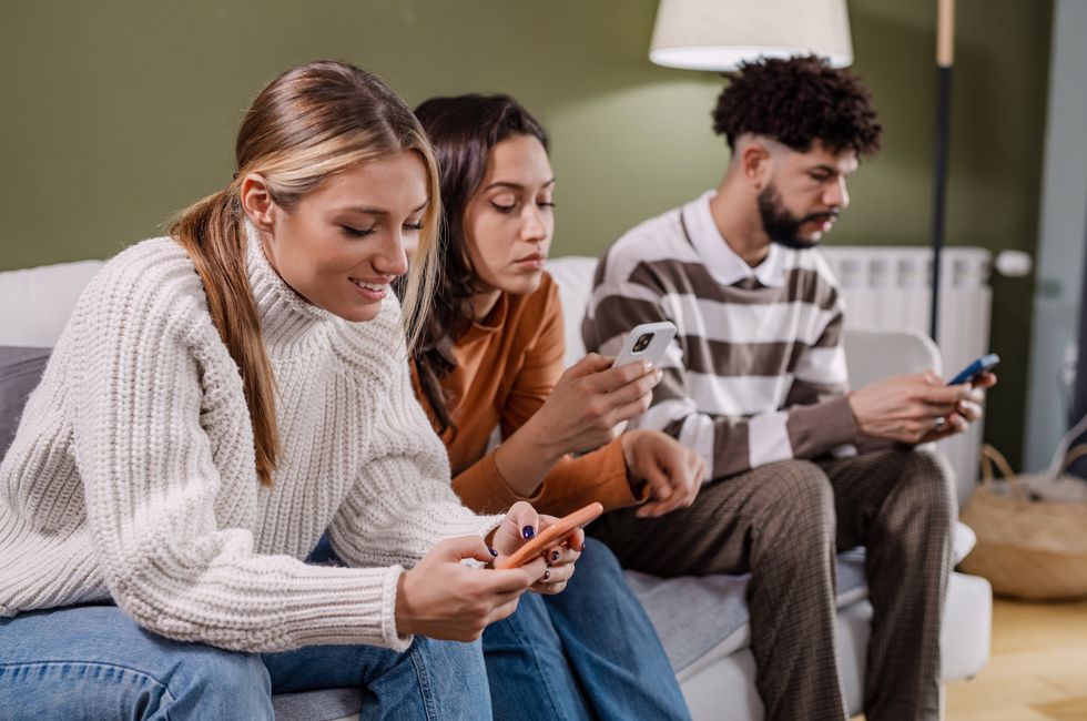 friends sitting on the couch in the living room and using mobile phones they are all looking at their screens while using fingers and thumbs to type
