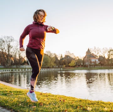 Checking heartbeat and pressure during jogging. Mature red haired female running near lake in green park female sportswoman in red sports wear monitoring and looking at wrist smartwatch screen