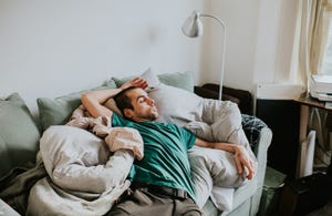 a man sleeps on top of duvets and pillows on a sofa in a living room