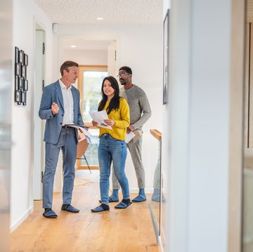 an adult caucasian male real estate agent giving an apartment tour to a young diverse couple the real estate agent is wearing a fancy suit while the couple is wearing casual clothes together they are walking through the hall of the apartment the rooms have modern decor with wooden details