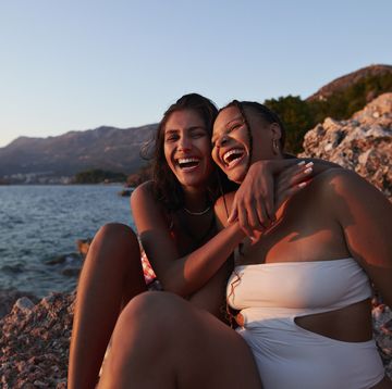 cheerful young woman sitting on rock with arm around friend enjoying sunset together at beach