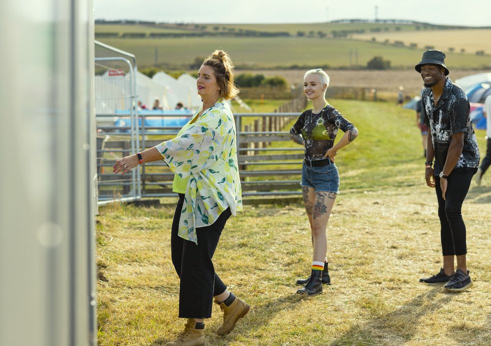 a group of festival goers at a festival in lindisfarne, north east england, standing in a field with the campsite in the background they are all queuing for the toilets and smiling as one woman is entering a cubicle
