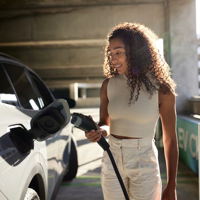 young woman with curly hair holding electric plug by car at charging station