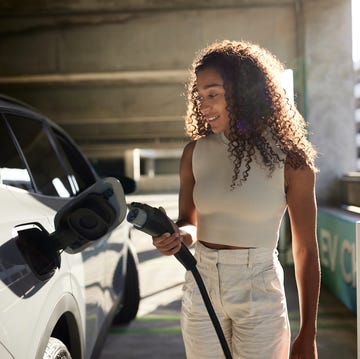 young woman with curly hair holding electric plug by car at charging station