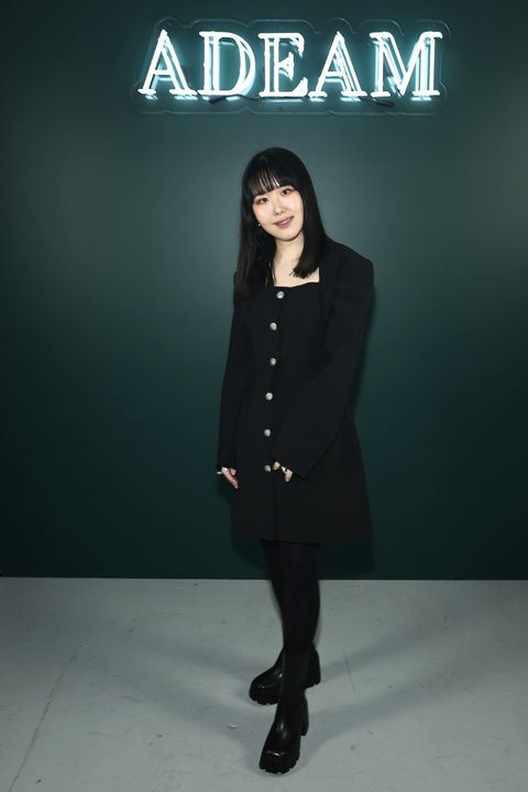 designer hanako maeda of adeam, in a short black dress with black tights and boots, stands in front of a neon sign that says adeam