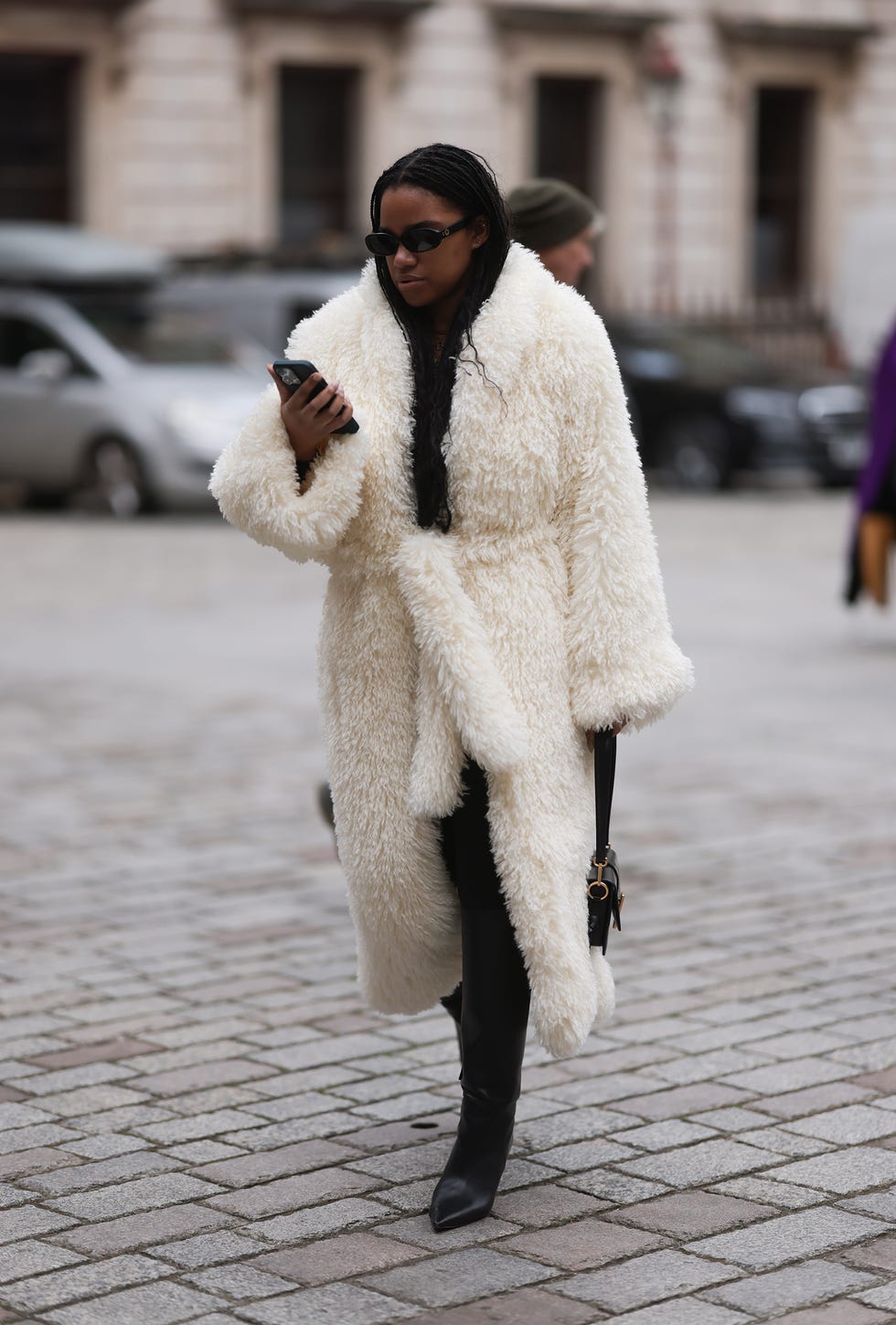 person wearing a long fluffy cream coat and black boots holding a phone while walking on a cobblestone street