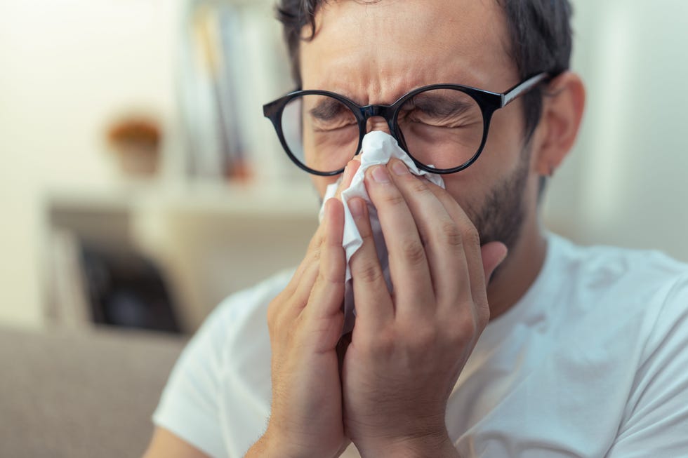 young man with glasses sneezing, wiping his nose with a piece of tissue paper