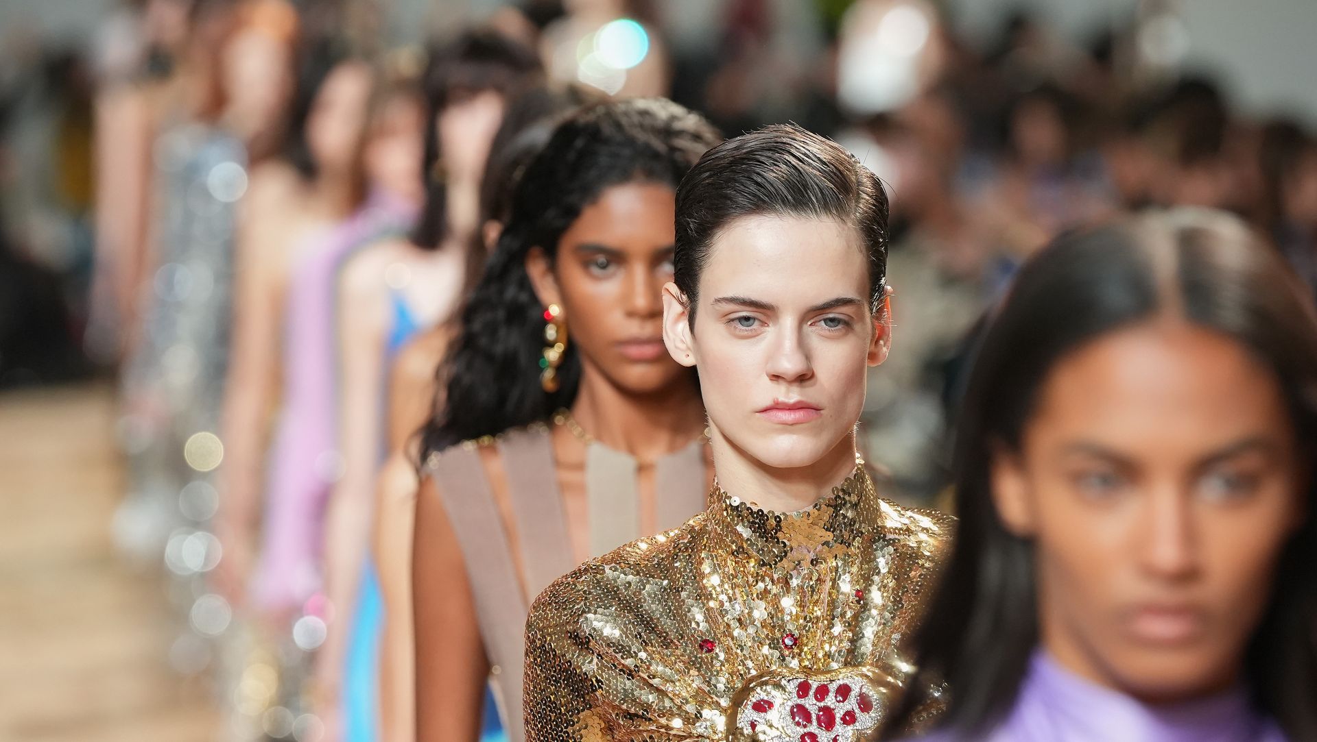 models walking in a line at the paco rabanne fall 2023 show in paris, with the camera focusing on one woman with a proud expression in a gold high necked dress