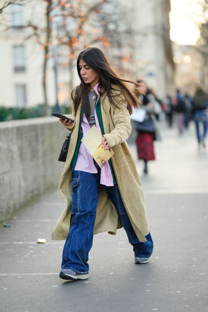 paris, france march 01 heloise salessy wears a gray t shirt, gold chain pendant necklaces, a pale pink and white striped print pattern shirt, a green zipper jacket, a beige pale yellow long coat, blue denim large cargo pants, a black shiny leather shoulder bag, gray sneakers , outside paco rabanne, during paris fashion week womenswear fall winter 2023 2024, on march 01, 2023 in paris, france photo by edward berthelotgetty images