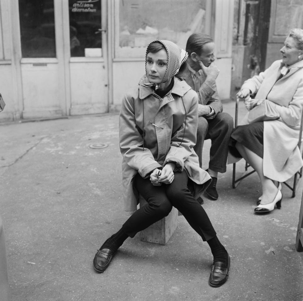 british actress audrey hepburn, wearing a raincoat and a headscarf, with american actor, dancer and singer fred astaire, and american singer, dancer and actress kay thompson sitting in the background, during a break in filming of funny face in paris, france, july 1956. directed by stanley donen, the musical features songs by george and ira gershwin. (photo by graphic house/archive photos/getty images)